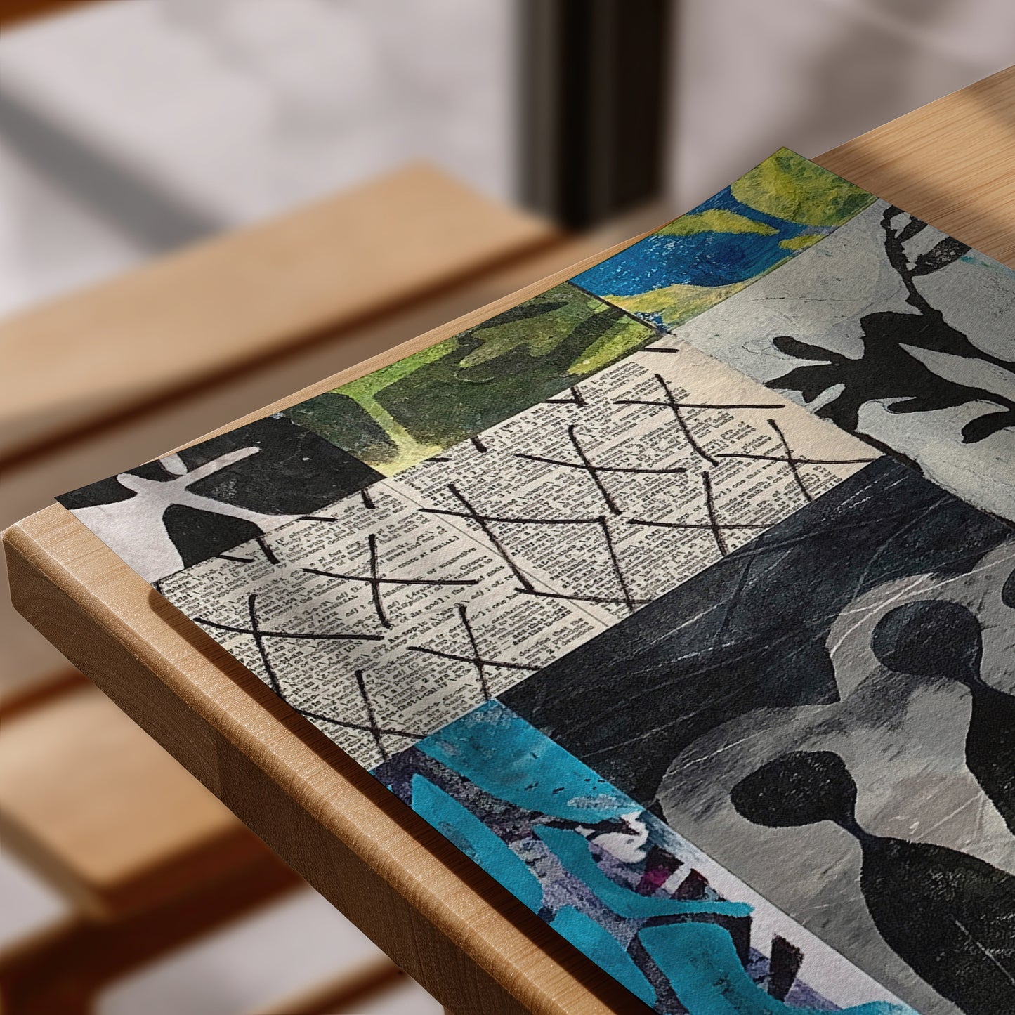 A close-up of a rectangular placemat featuring an abstract collage design with newspaper print, black ink sketches, and blue and green accents, resting on a light wood table.