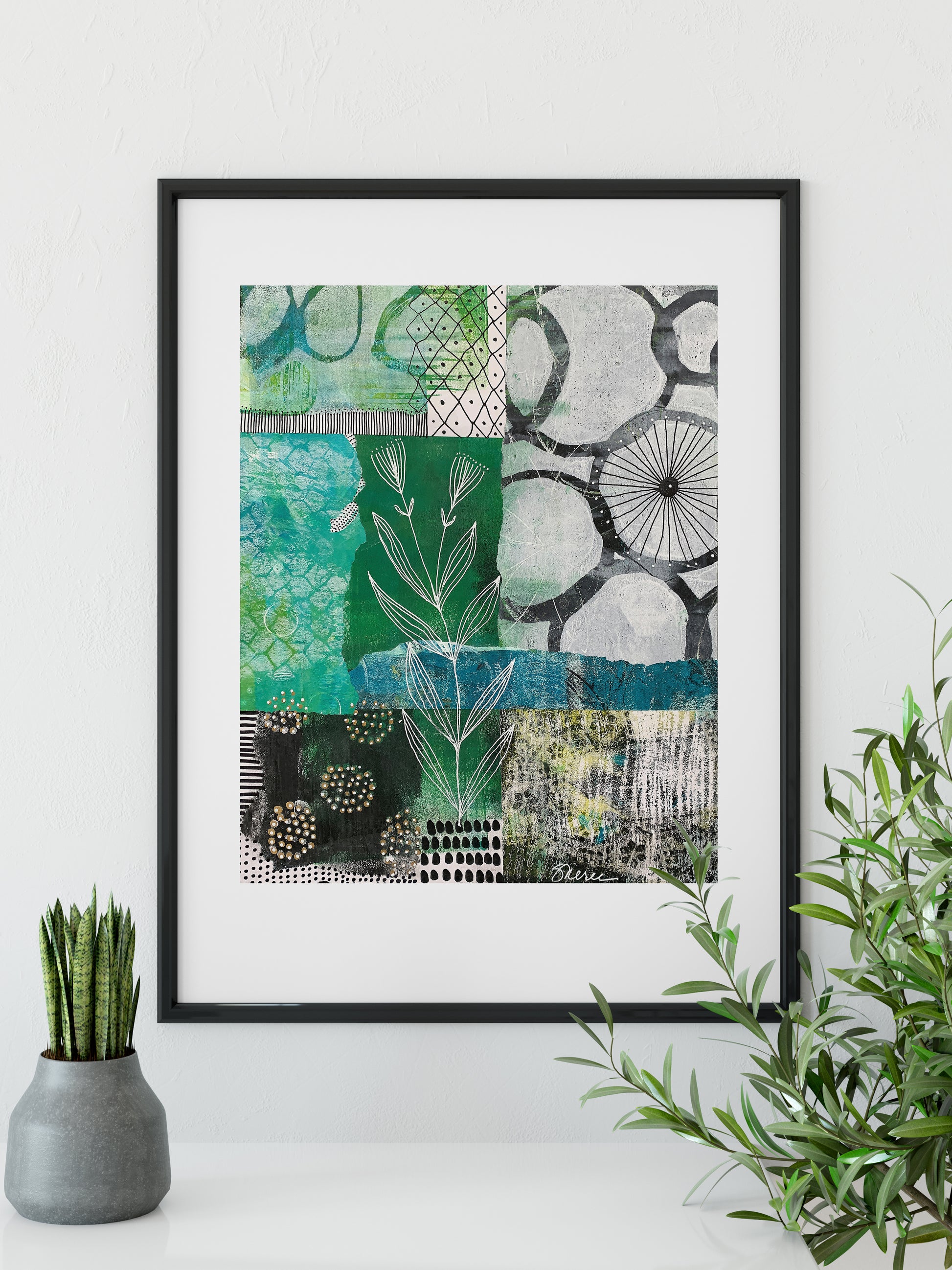 A framed abstract art print with green, teal, black, and white patterns, displayed on a white surface with a small potted succulent on the left and a green plant on the right.