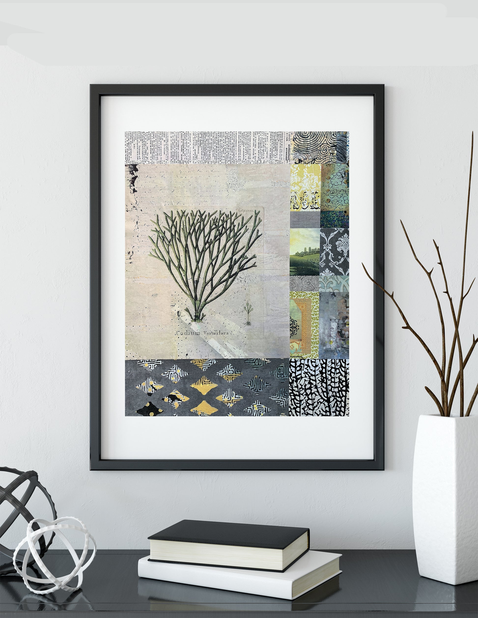Framed abstract patchwork art print featuring a coral-like silhouette and geometric patterns in muted gray, beige, black, yellow, and green tones, displayed on a shelf above stacked books with decorative branches in a white vase.