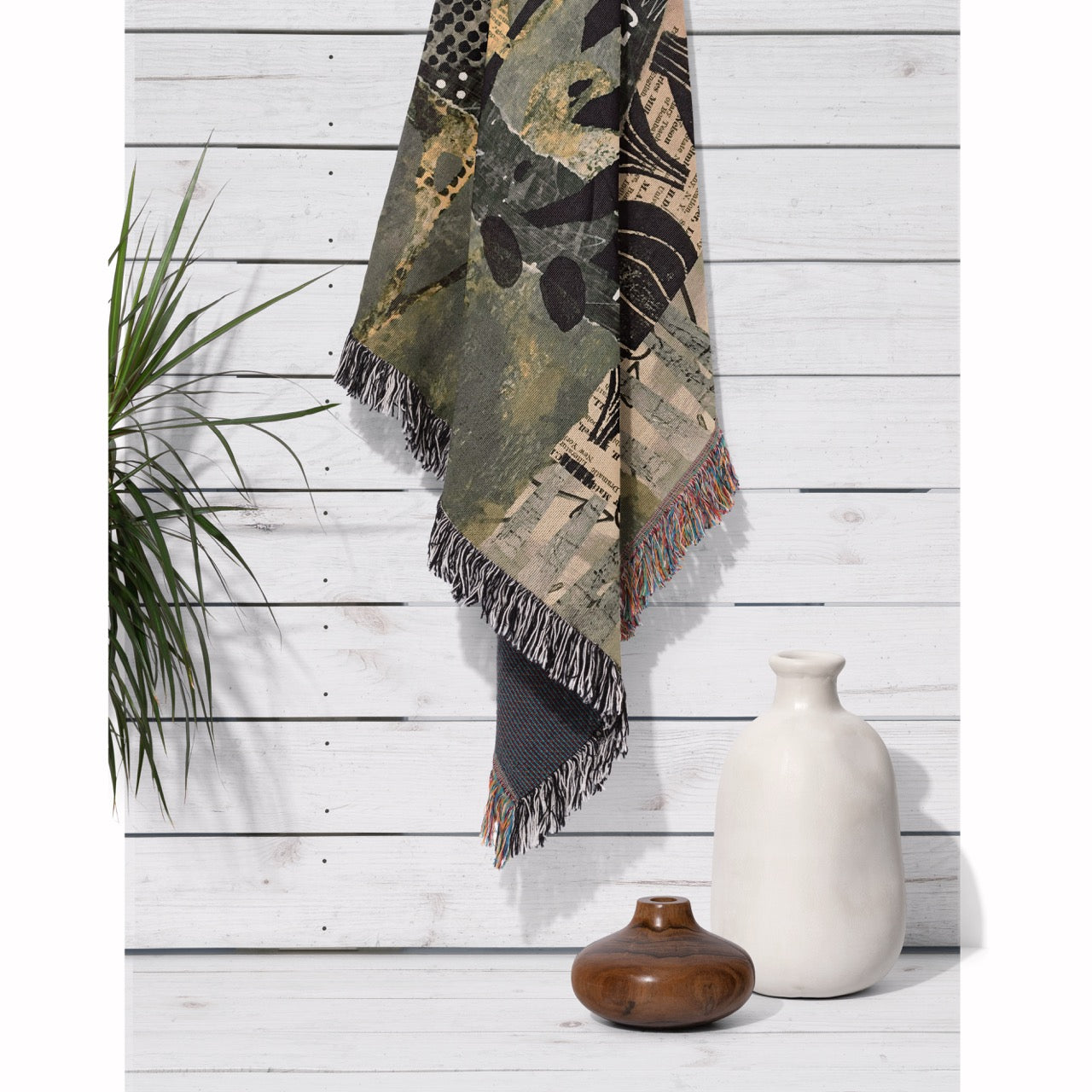 Decorative blanket with fringe hanging on a wooden wall with vases and a plant.
